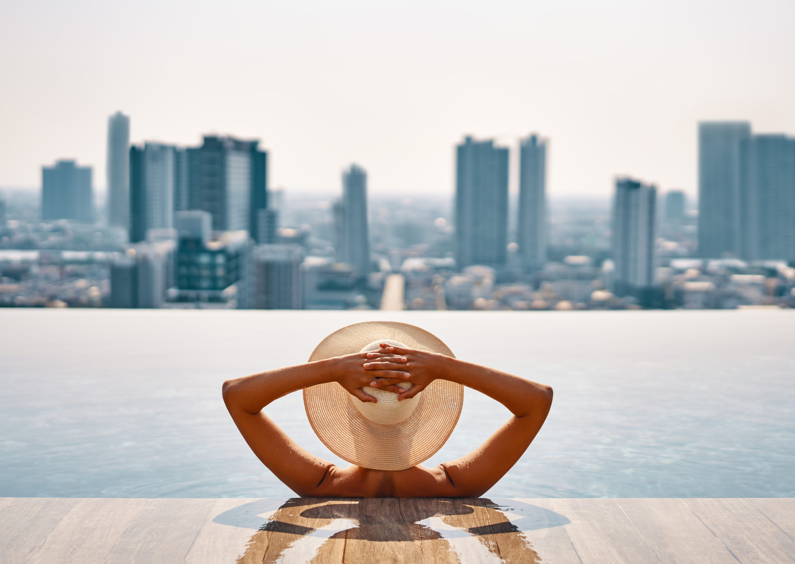 Back view of young woman in hat relaxing in swimming pool on the roof top of hotel and enjoy cityscape